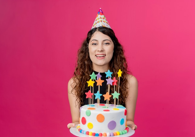 Young beautiful woman with curly hair in a holiday cap holding birthday cake hapy and cheerful birthday party concept  over pink