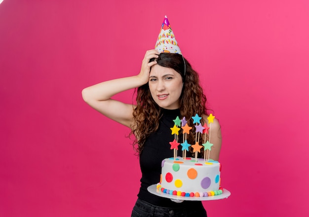 Молодая красивая женщина с вьющимися волосами в праздничной шапочке держит торт ко дню рождения, смущенный рукой на голове, концепция вечеринки по случаю дня рождения, стоящая над розовой стеной