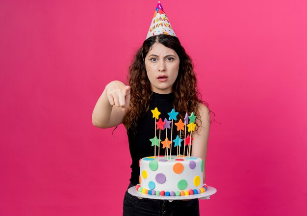 ピンクの壁の上に立っている指で不愉快な誕生日パーティーのコンセプトを指しているバースデーケーキを保持しているホリデーキャップの巻き毛の若い美しい女性