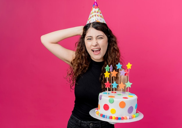 생일 케이크를 들고 휴가 모자에 곱슬 머리를 가진 젊은 아름 다운 여자 핑크 벽 위에 서 행복하고 즐거운 생일 파티 개념을 유쾌하게 찾고 웃고
