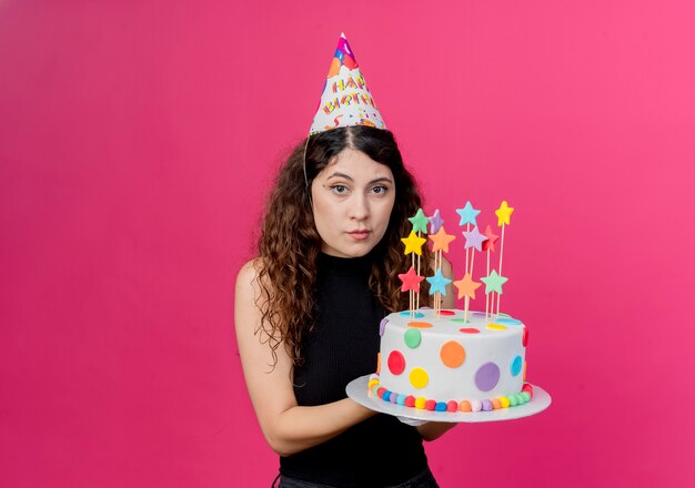 Young beautiful woman with curly hair in a holiday cap holding birthday cake  with serious face birthday party concept standing over pink wall