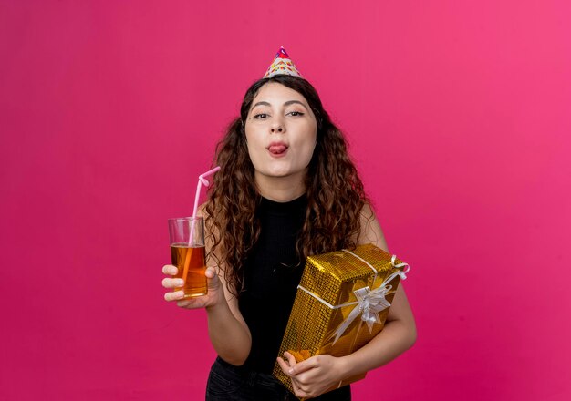 Young beautiful woman with curly hair in a holiday cap holding birthday gift box and cocktail looking at front happy and positive sticking out tongue birthday party