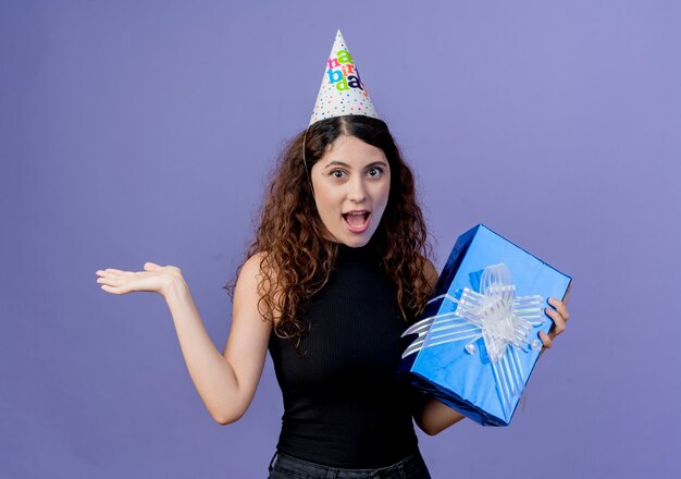 Молодая красивая женщина с вьющимися волосами в праздничной кепке держит подарочную коробку на день рождения, выглядит изумленной и удивленной концепции вечеринки по случаю дня рождения, стоящей над синей стеной