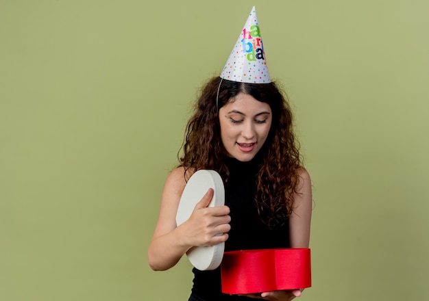 Free photo young beautiful woman with curly hair in a holiday cap holding gift box looking at it surprised and happy birthday party concept  over light