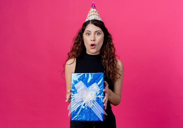 Young beautiful woman with curly hair in a holiday cap holding gift box looking surprised and amazed birthday party concept standing over pink wall