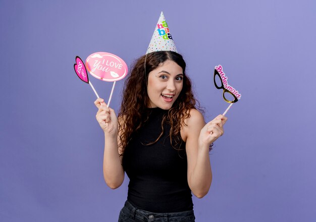 Young beautiful woman with curly hair in a holiday cap holding holding holiday mask smiling cheerfully birthday party concept standing over blue wall