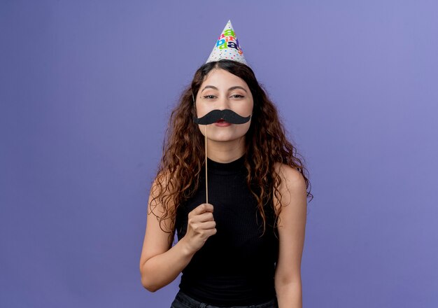 Young beautiful woman with curly hair in holiday cap holding mustache party sticks standing over blue wall