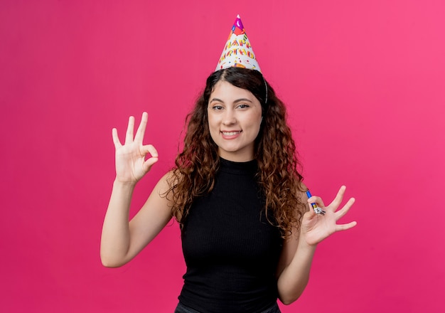 Молодая красивая женщина с вьющимися волосами в праздничной шапочке держит свисток, показывая знак ОК, весело улыбаясь, концепция вечеринки по случаю дня рождения, стоя над розовой стеной