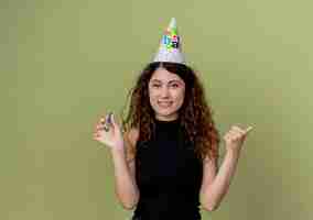 Free photo young beautiful woman with curly hair in a holiday cap holding whistle smiling cheerfully birthday party concept standing over light wall