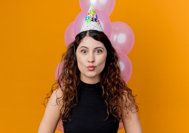 Young beautiful woman with curly hair in a holiday cap looking at canera surprised birthday party concept standing with balloons over orange wall