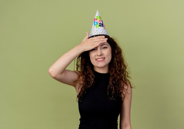 Young beautiful woman with curly hair in a holiday cap looking confused smiling with hand over head birthday party concept standing over orange wall