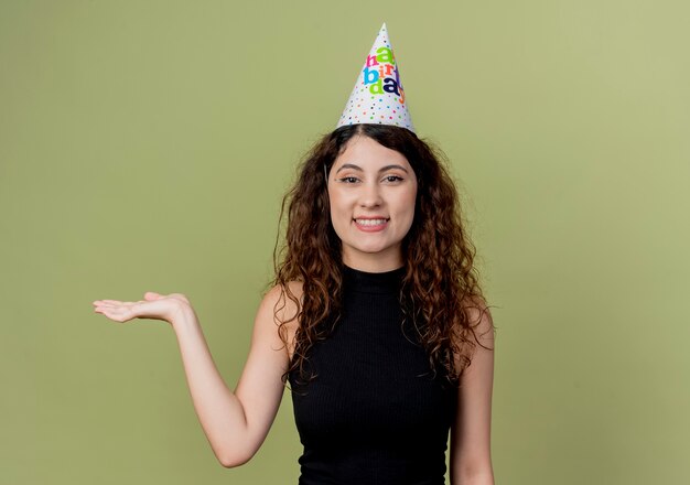 Молодая красивая женщина с вьющимися волосами в праздничной кепке, представляя рукой, весело улыбаясь концепции вечеринки по случаю дня рождения, стоя над светлой стеной