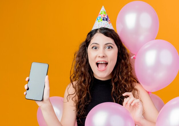 Молодая красивая женщина с вьющимися волосами в праздничной кепке показывает смартфон счастливой и взволнованной концепцией вечеринки по случаю дня рождения, стоящей с воздушными шарами над оранжевой стеной