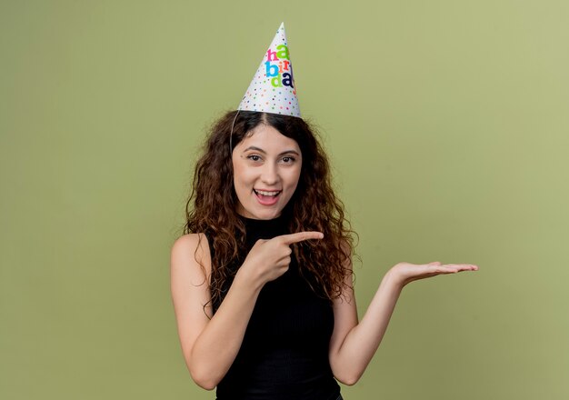 Young beautiful woman with curly hair in a holiday cap smiling pointing with finger to the side presenting with arm of hand birthday party concept  over light
