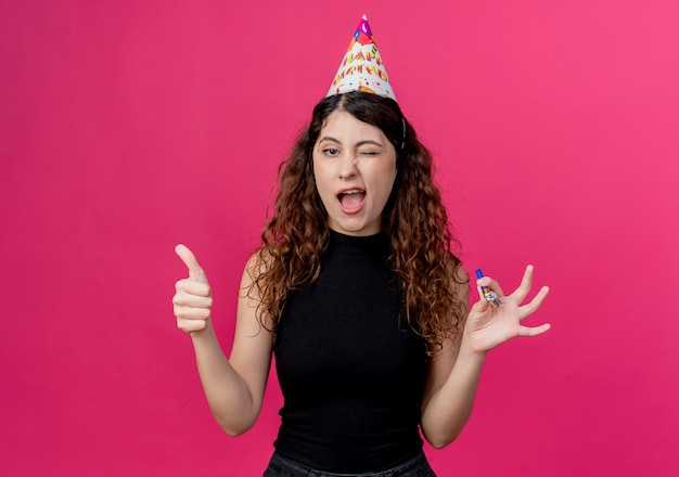Giovane bella donna con capelli ricci in un berretto da vacanza sorridente e ammiccante che mostra i pollici in su concetto di festa di compleanno in piedi sopra la parete rosa
