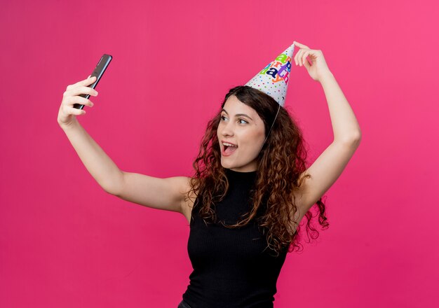 ピンクの上の誕生日パーティーのコンセプトを元気に笑顔で自分撮りを取っているホリデーキャップの巻き毛の若い美しい女性