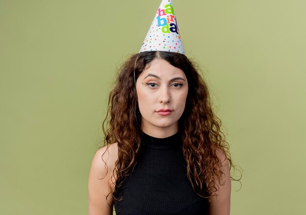 Young beautiful woman with curly hair in a holiday cap with sad expression birthday party concept  over light