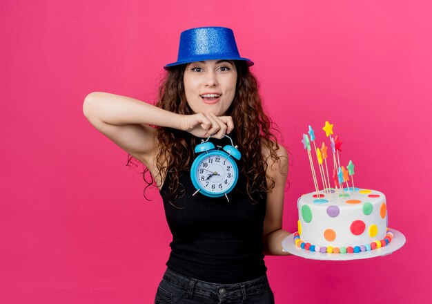 Young beautiful woman with curly hair in a holiday hat holding birthday cake and alarm clock looking surprised and happy birthday party concept standing over pink wall