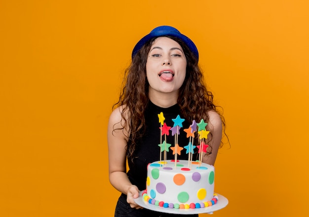 Young beautiful woman with curly hair in a holiday hat holding birthday cake crazy happy sticking out tongue standing over orange wall