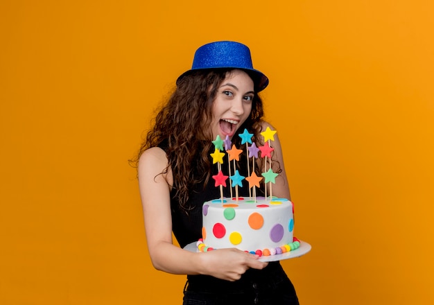 Foto gratuita giovane bella donna con capelli ricci in un cappello di festa che tiene la torta di compleanno felice e allegra in piedi sopra la parete arancione
