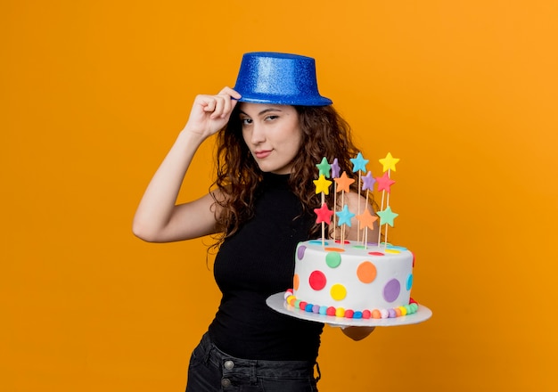 Young beautiful woman with curly hair in a holiday hat holding birthday cake  smiling confident standing over orange wall