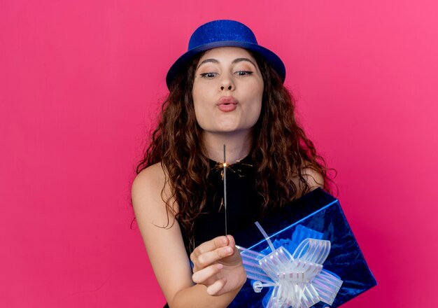 Young beautiful woman with curly hair in a holiday hat holding birthday gift box and blowing on a sparkler birthday party concept standing over pink wall