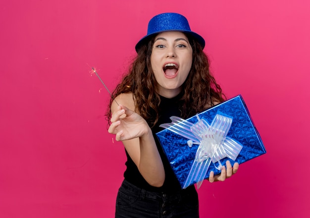 Young beautiful woman with curly hair in a holiday hat holding birthday gift box and sparkler happy and excited birthday party concept over pink