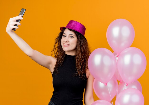 Young beautiful woman with curly hair in a holiday hat holding bunch of air balloons doing selfie smiling cheerfully birthday party concept standing over orange wall