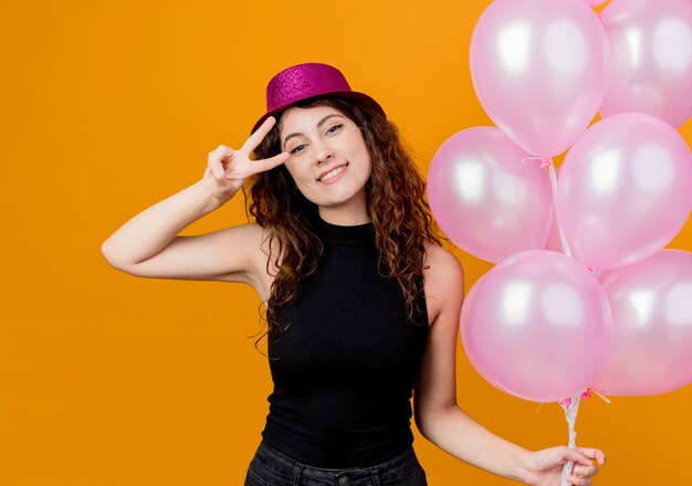 Молодая красивая женщина с вьющимися волосами в праздничной шляпе, держащая кучу воздушных шаров, счастлива и позитивно улыбается, весело показывая концепцию вечеринки по случаю дня рождения v-sign, стоящую над оранжевой стеной