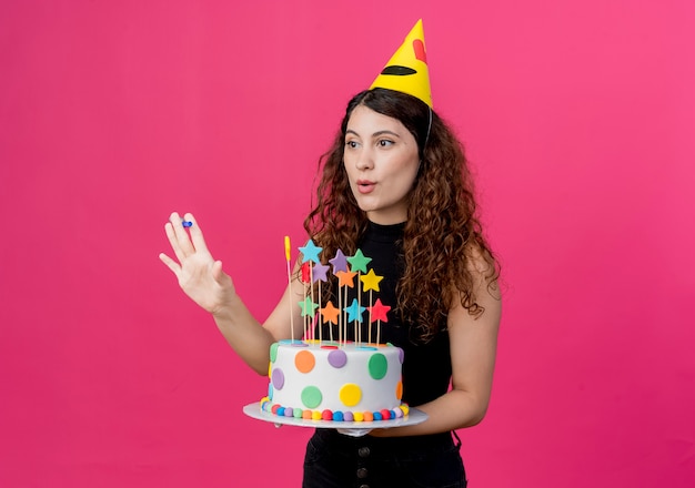 Молодая красивая женщина с вьющимися волосами в праздничной шапочке держит праздничный торт с счастливой и позитивной концепцией вечеринки по случаю дня рождения, стоящей над розовой стеной