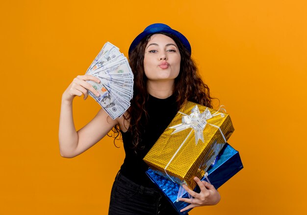 Young beautiful woman with curly hair in party hat holding cash and gifts looking at canera happy and cheerful blowing a kiss over orange