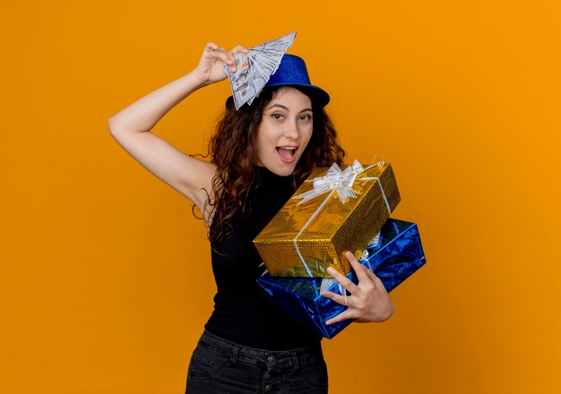 Young beautiful woman with curly hair in party hat holding cash and gifts looking at canera happy and excited standing over orange wall