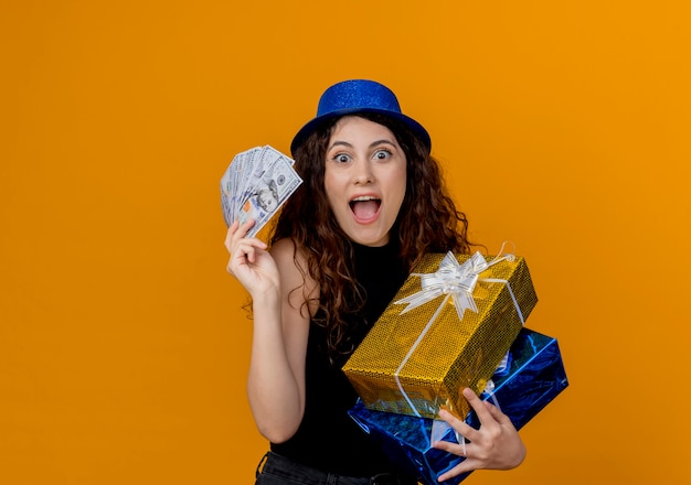 Молодая красивая женщина с вьющимися волосами в партийной шляпе держит деньги и подарки, глядя на счастливую и взволнованную канеру, стоящую над оранжевой стеной