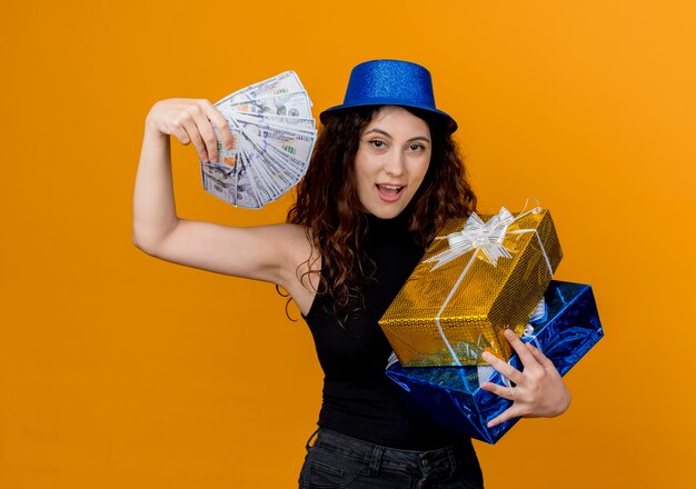 Free photo young beautiful woman with curly hair in party hat holding cash and gifts looking at canera happy and excited standing over orange wall