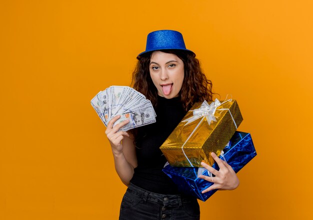 Young beautiful woman with curly hair in party hat holding cash and gifts looking at canera happy and excited sticking out tongue over orange