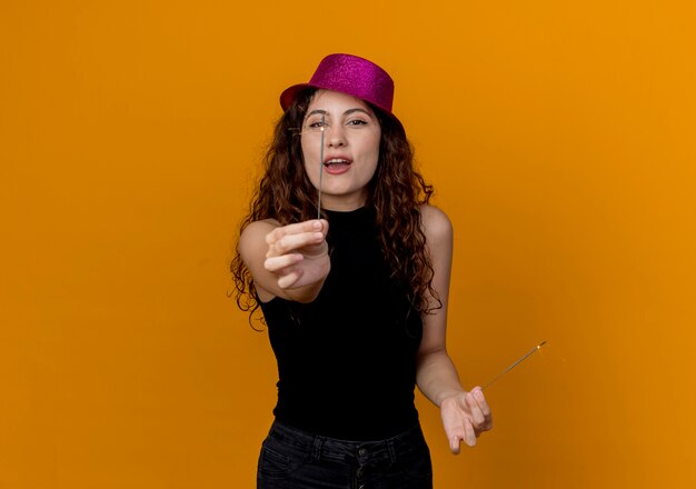 Young beautiful woman with curly hair in party hat holding sparklers  happy and excited standing over orange wall