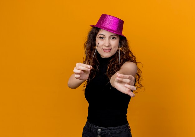 Giovane bella donna con capelli ricci in cappello del partito che tiene le stelle filanti felici ed eccitate in piedi sopra la parete arancione