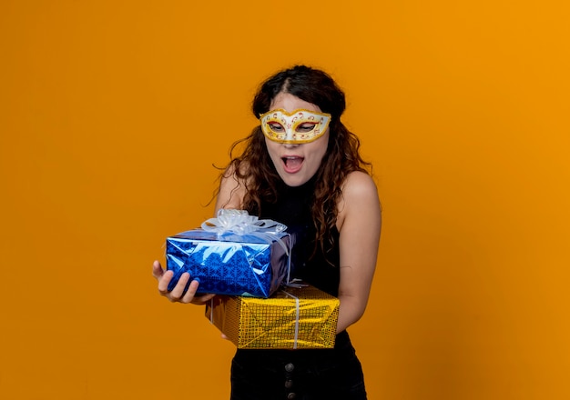 Молодая красивая женщина с вьющимися волосами в маске для вечеринки держит счастливые и взволнованные подарки, стоя над оранжевой стеной