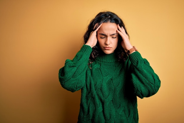 무료 사진 노란색 배경 위에 녹색 캐주얼 스웨터를 입은 곱슬머리의 젊은 미녀 두통과 편두통으로 고통받고 스트레스를 받고 있습니다.