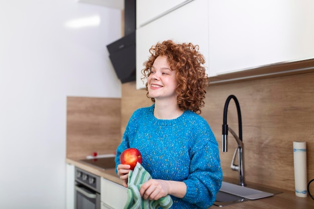Бесплатное фото Молодая красивая женщина с вьющимися рыжими волосами ест сочное красное яблоко, стоя дома на кухне ежедневное потребление витаминов с фруктами диета и здоровое питание