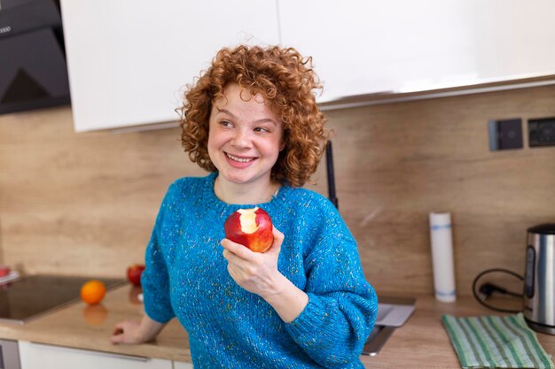 Young beautiful woman with curly red hair eating a juicy red apple while standing in her kitchen at home Daily intake of vitamins with fruits Diet and healthy eating