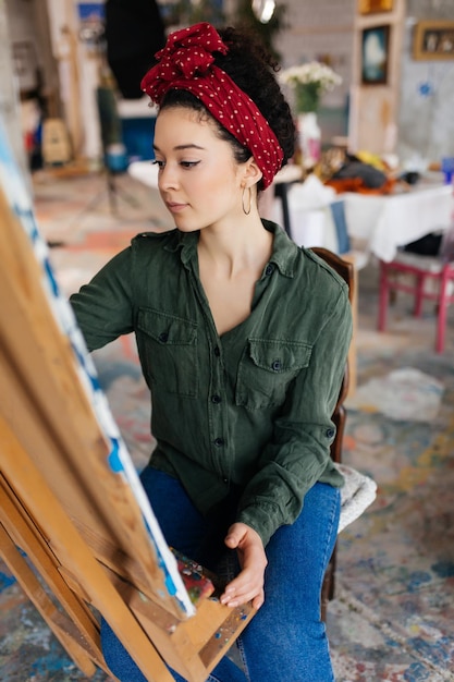 Foto gratuita giovane bella donna con capelli ricci scuri seduto su una sedia che disegna sognante un'immagine su tela mentre trascorre del tempo in un moderno laboratorio d'arte accogliente