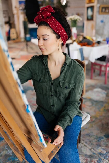 Молодая красивая женщина с темными вьющимися волосами сидит на стуле, мечтательно рисуя картину на холсте, проводя время в современной уютной художественной мастерской