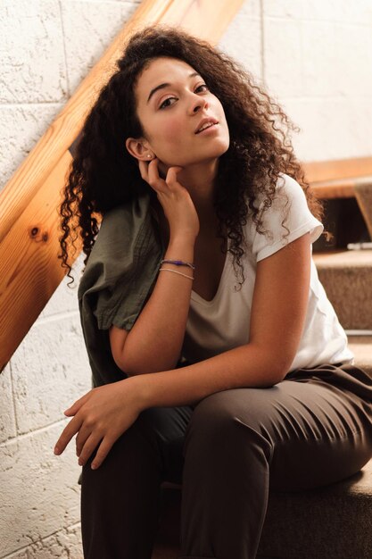 Young beautiful woman with dark curly hair sitting on stairs in university and dreamily looking in camera