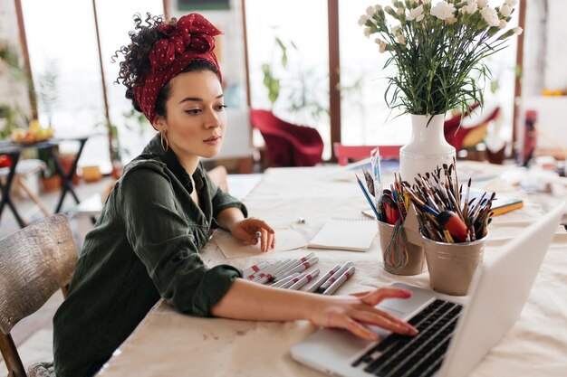 Young beautiful woman with dark curly hair sitting at the table dreamily using laptop and drawing sketches spending time in modern cozy workshop with big windows