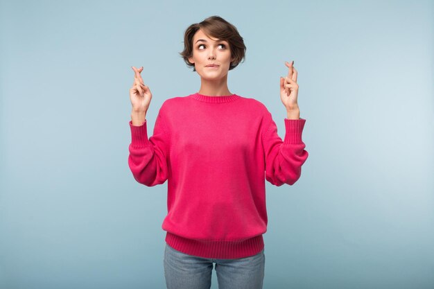 Young beautiful woman with dark short hair in pink sweater keeping fingers crossed while dreamily looking aside over blue background