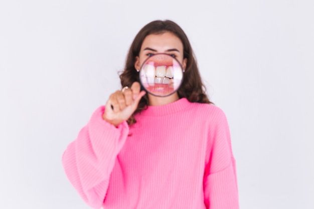 Young beautiful woman with freckles light makeup in sweater on white wall with magnifier  shows white teeth perfect smile