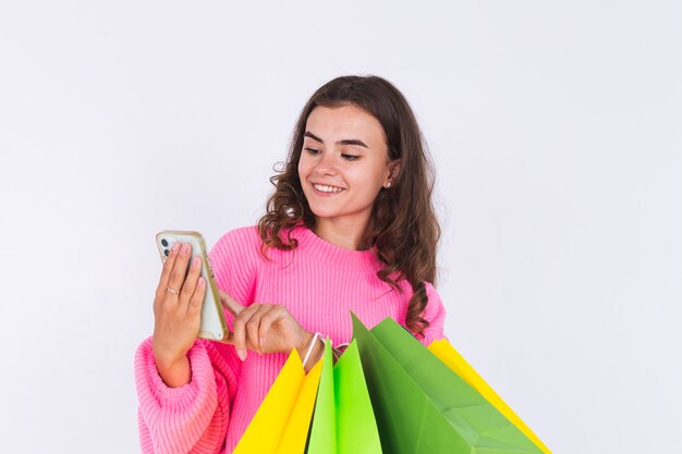 そばかすのある若い美しい女性は、買い物袋と携帯電話の笑顔で陽気なポジティブな白い壁にセーターで軽い化粧をします。