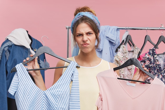 Young beautiful woman with grumpy expression, frowning her face with dissatisfaction while holding two dresses, having no suitable size for her. Discontent woman having problems during shopping