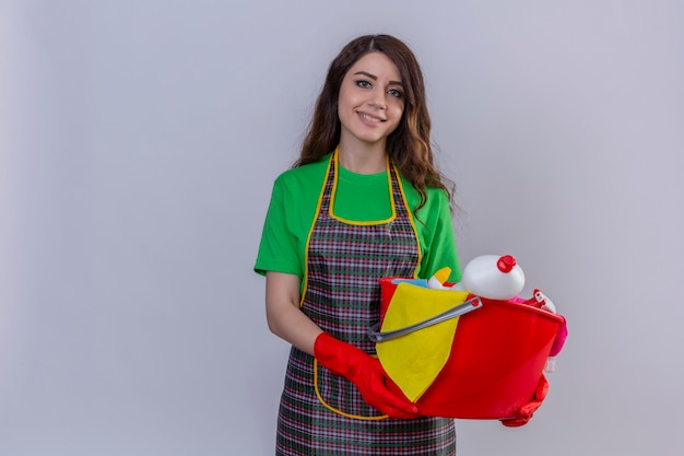 Young beautiful woman with long wavy hair in apron and gloves holding bucket with cleaning tools smiling friendly standing 